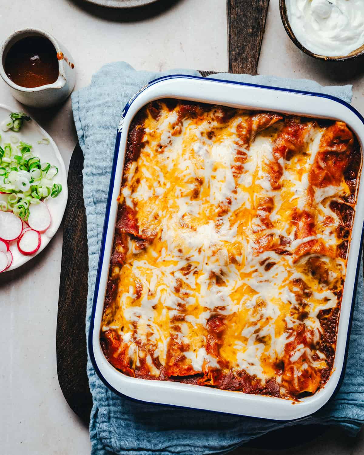 A baked dish of cheesy enchiladas in a white and blue casserole dish. The top is golden with melted cheese. Surrounding the dish are a container of sauce, a bowl of sour cream, and a plate with sliced radishes and green onions.