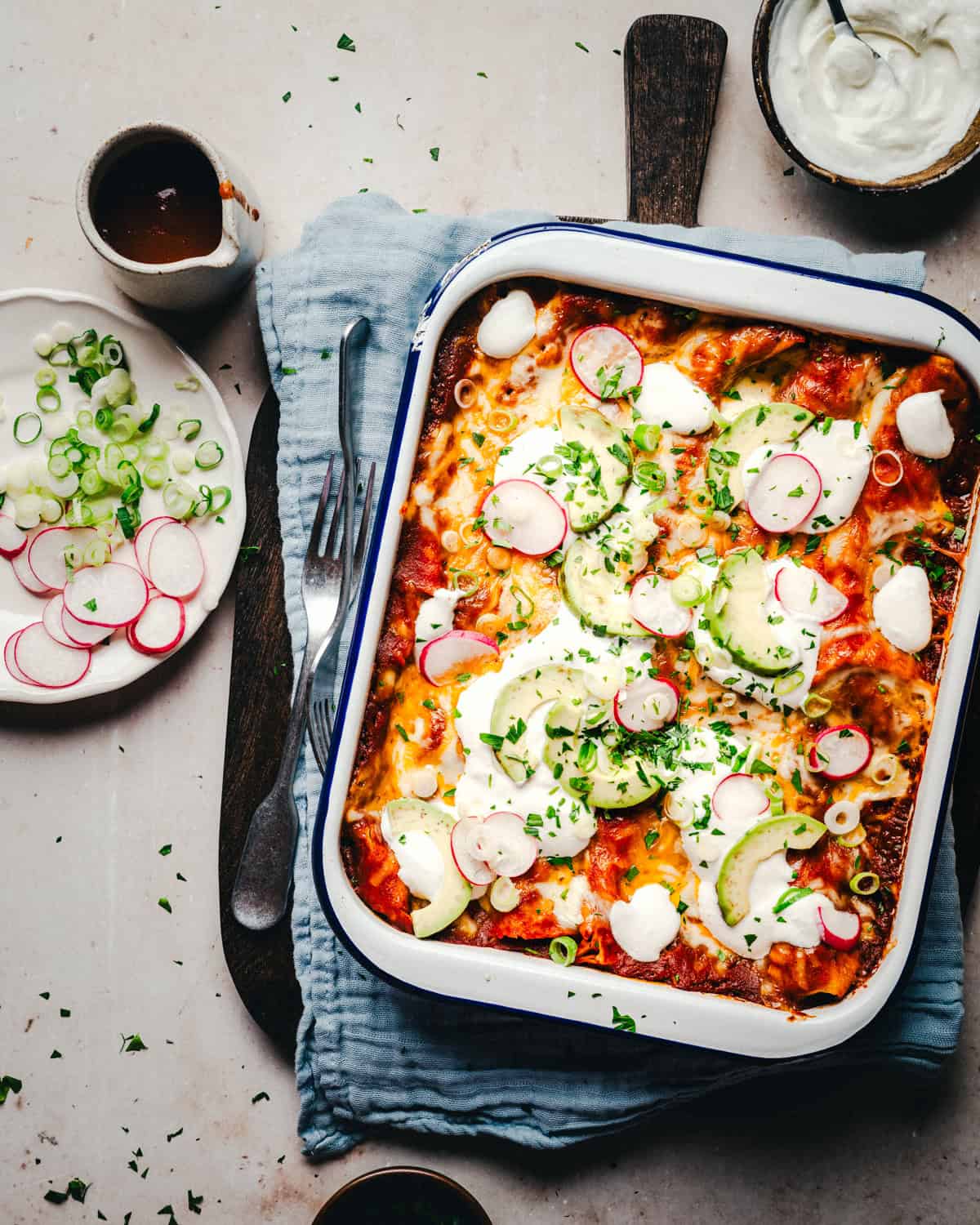 A baked cheese enchilada topped with sliced avocados, radishes, green onions, and dollops of sour cream. Nearby, a plate holds extra radish and green onion slices. A small bowl of sauce and a fork rest on a cloth beneath the dish.