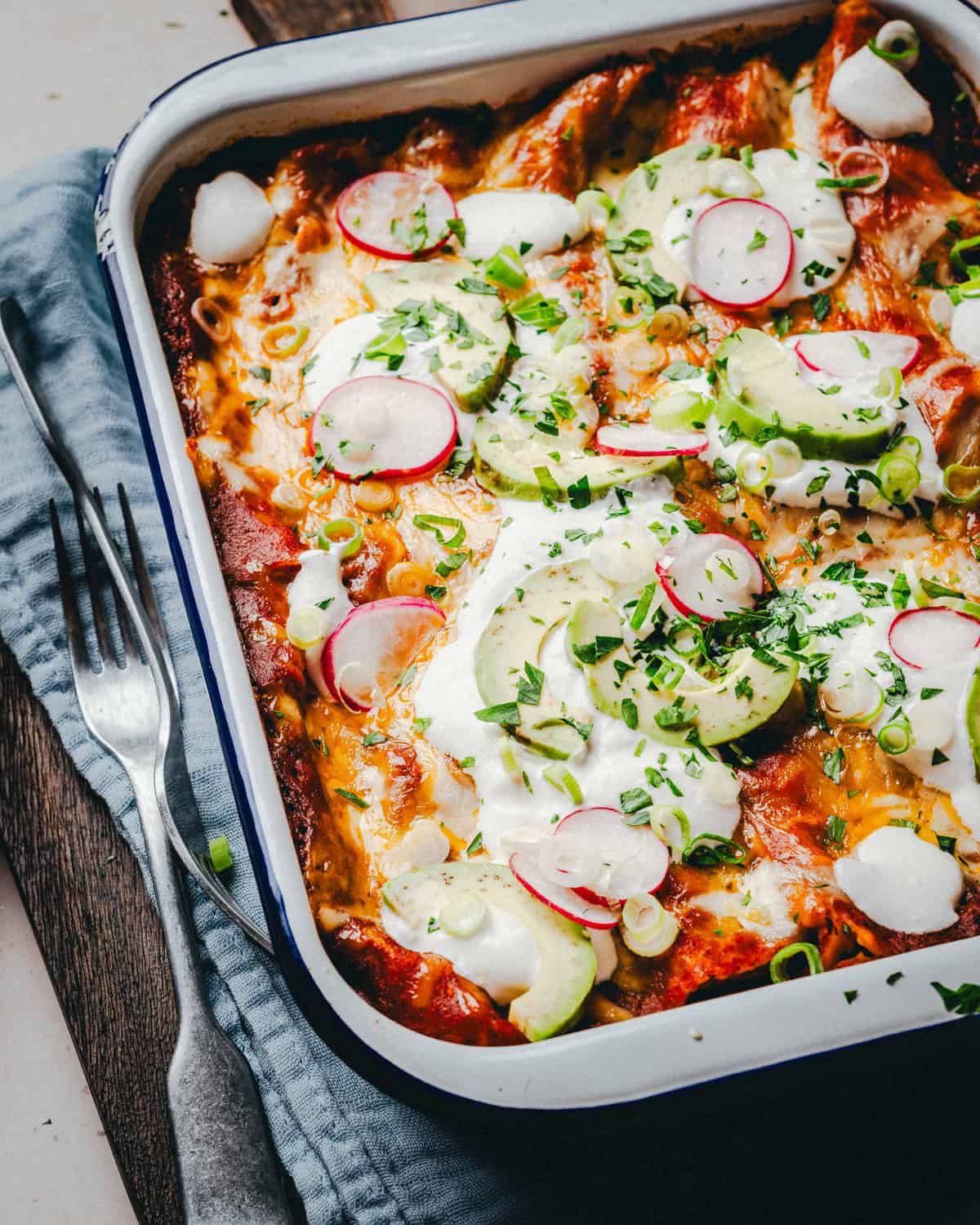 A baked dish of cheese enchiladas topped with sour cream, sliced radishes, avocado, and chopped green onions. The dish is in a white rectangular baking tray on a dark wooden surface with a fork and knife on a gray cloth beside it.