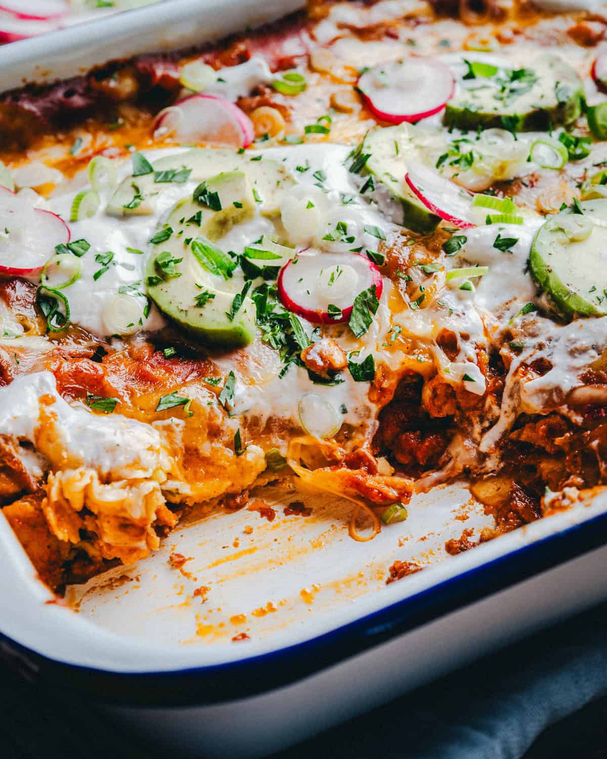Cheese enchiladas with a cheesy top layer, garnished with sliced radishes, avocados, and green onions. The dish has a missing portion, revealing layers of tortilla, cheese and sauce. It is served in a rectangular white dish.