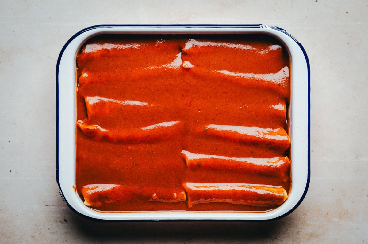 A white and blue-rimmed rectangular dish filled with several rolled tortillas covered in a rich, red enchilada sauce, ready for baking. The dish is set on a light beige surface.