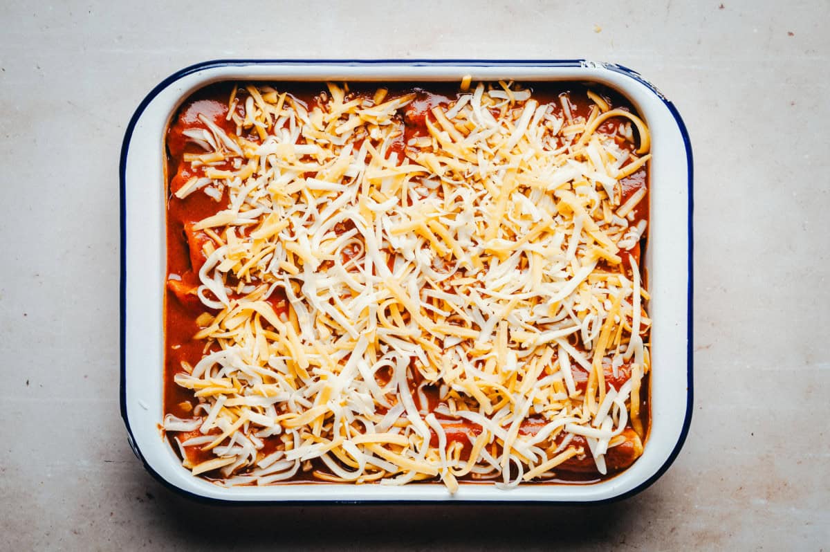 A rectangular baking dish filled with enchiladas, topped with red sauce and a generous layer of shredded cheese, placed on a light-colored surface.