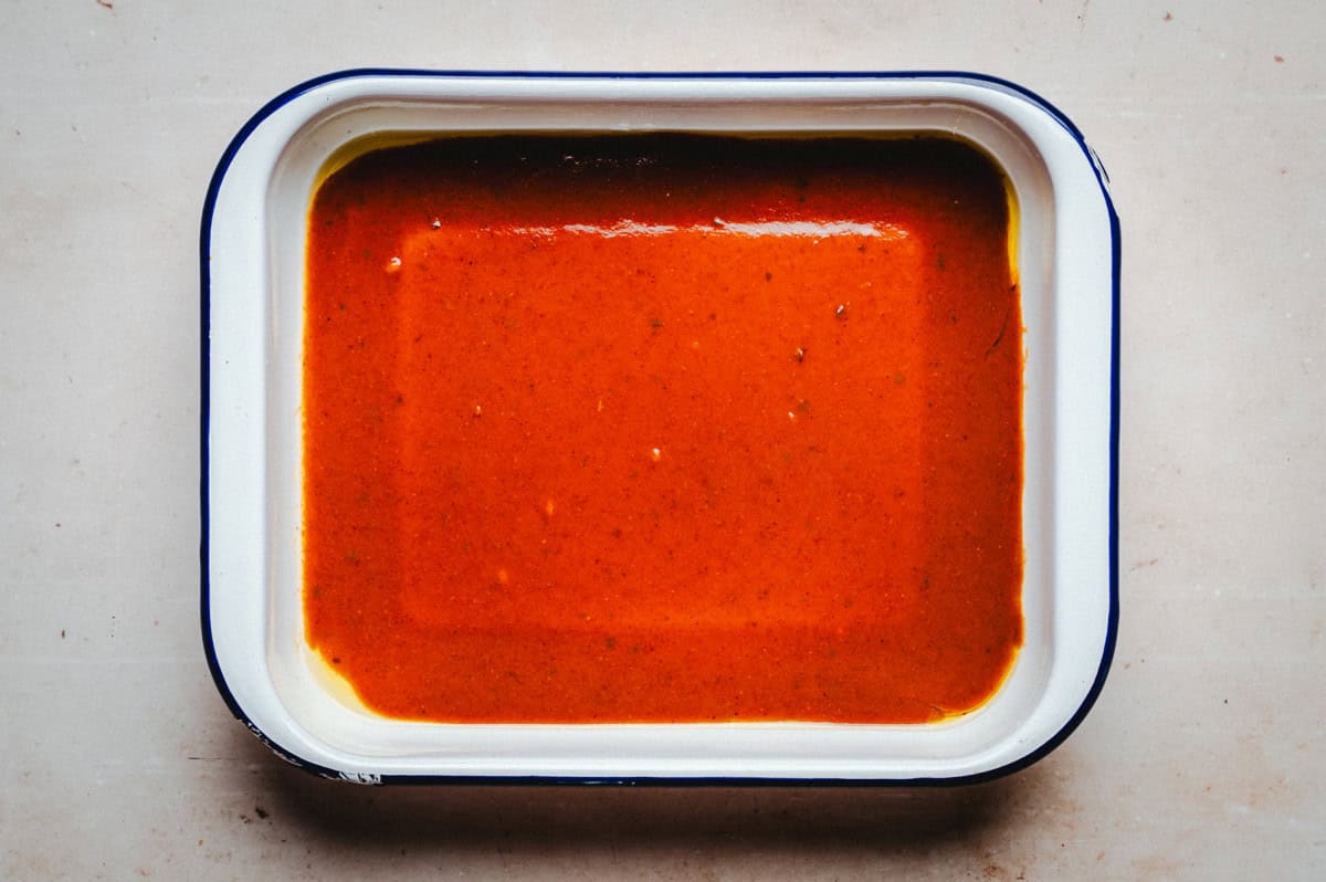 A rectangular white baking dish with a blue rim filled with bright red sauce. The sauce has a smooth texture with a slightly glossy surface. The dish is placed on a light-colored countertop.