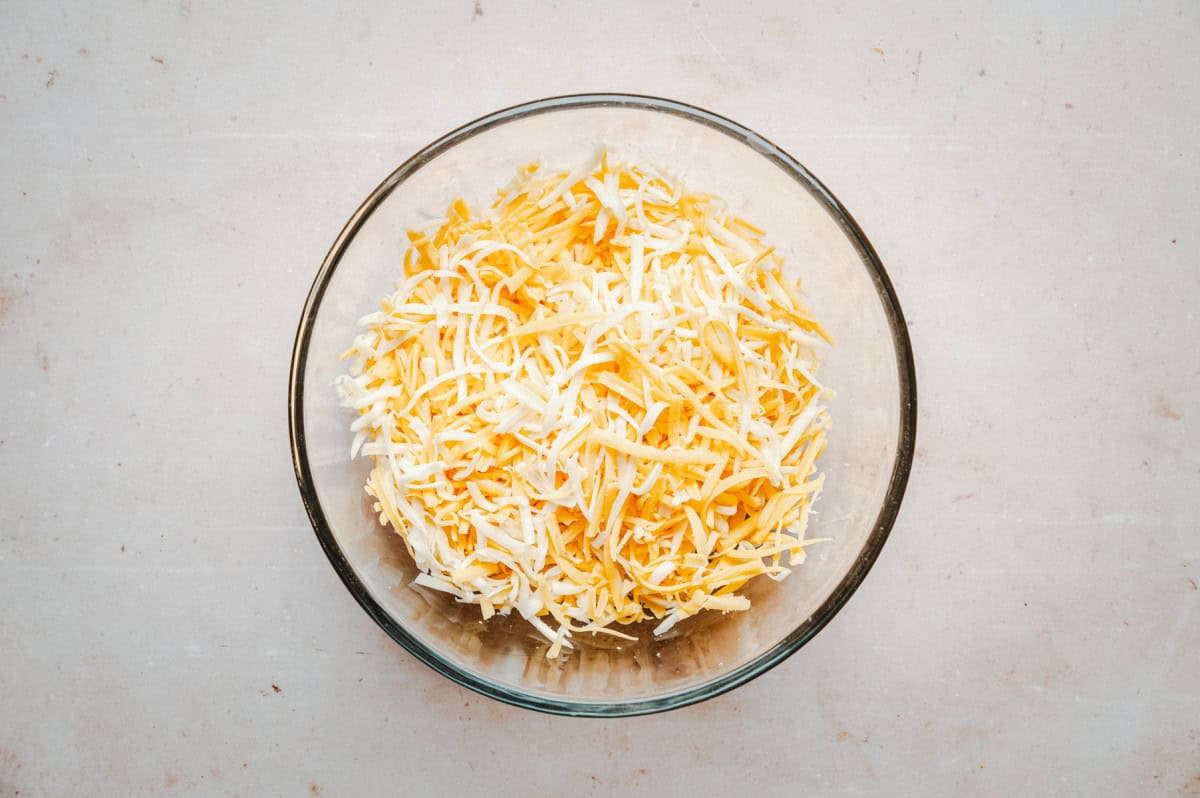 A glass bowl filled with shredded cheddar and jack cheese, placed on a light-colored, textured surface.