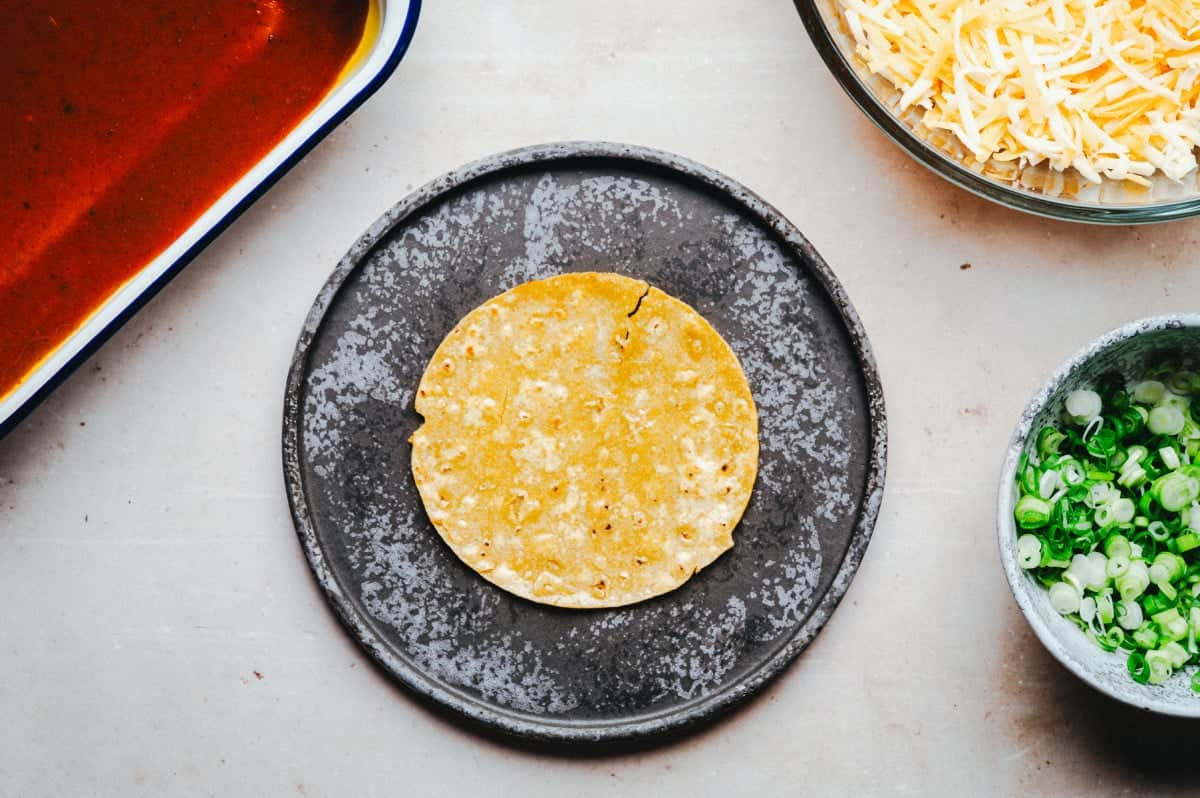 A corn tortilla on a dark plate, surrounded by a bowl of shredded cheese, a bowl of chopped green onions, and a baking dish with red sauce, set on a light surface.