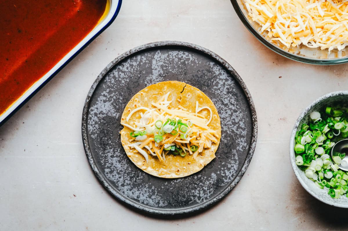 A corn tortilla with shredded cheese and chopped green onions is on a dark plate. Nearby are bowls with more shredded cheese, chopped green onions, and sauce. The background is a light surface.