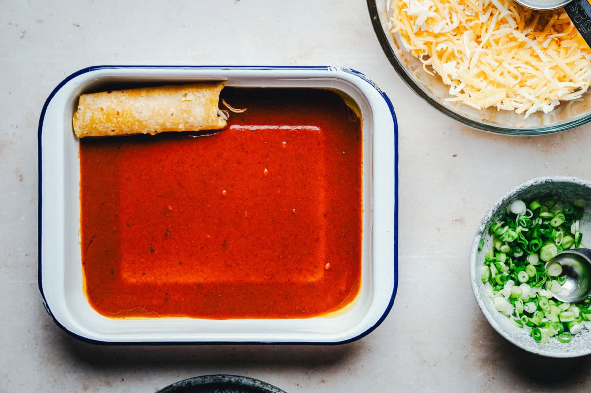 A baking dish with an enchilada in red sauce, positioned diagonally. Nearby, there are bowls with shredded cheese and sliced green onions on a light-colored surface.
