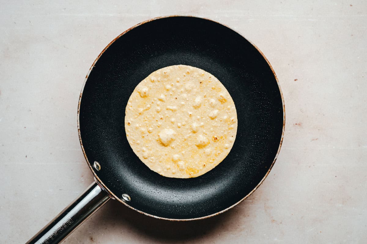 A single tortilla cooking in a black non-stick frying pan on a light beige surface. The tortilla is golden-brown with small bubbles, centered in the pan.