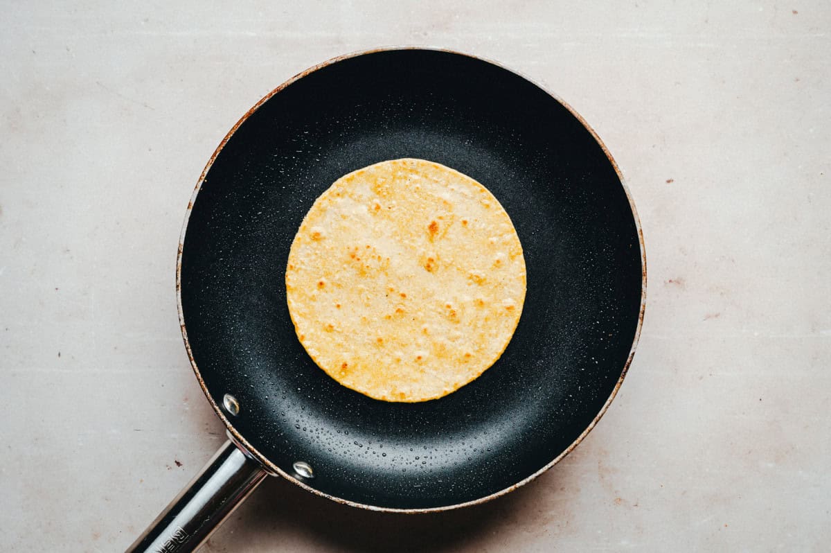 A single corn tortilla is being heated in a black frying pan on a light-colored surface.