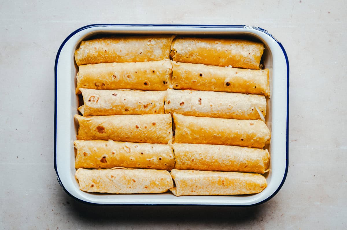 A white rectangular baking dish filled with neatly arranged, rolled tortillas. They are golden-brown and are tightly packed in two rows. The background is a light, neutral-colored surface.