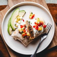 A chorizo burrito topped with diced pineapple, red peppers, and white sauce sits on a white plate. Slices of avocado are on the side. A fork rests on the plate, and the dish is placed on a wooden board.
