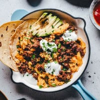 A skillet filled with scrambled eggs topped with sour cream, Mexican chorizo sausage, and chopped herbs. Sliced avocado and folded tortillas are on the side. A small bowl with red sauce is visible at the corner.