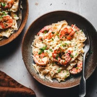 A bowl of creamy pasta with shrimp garnished with chopped parsley. A fork rests on the right side of the bowl, and part of another similar dish is visible in the top left corner. A brown napkin is nearby.