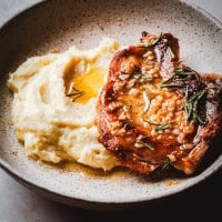A bowl featuring a seared pork chop with rosemary and garlic on top, served alongside creamy mashed potatoes with a small pool of melted butter. The dish is presented in a speckled brown ceramic bowl.