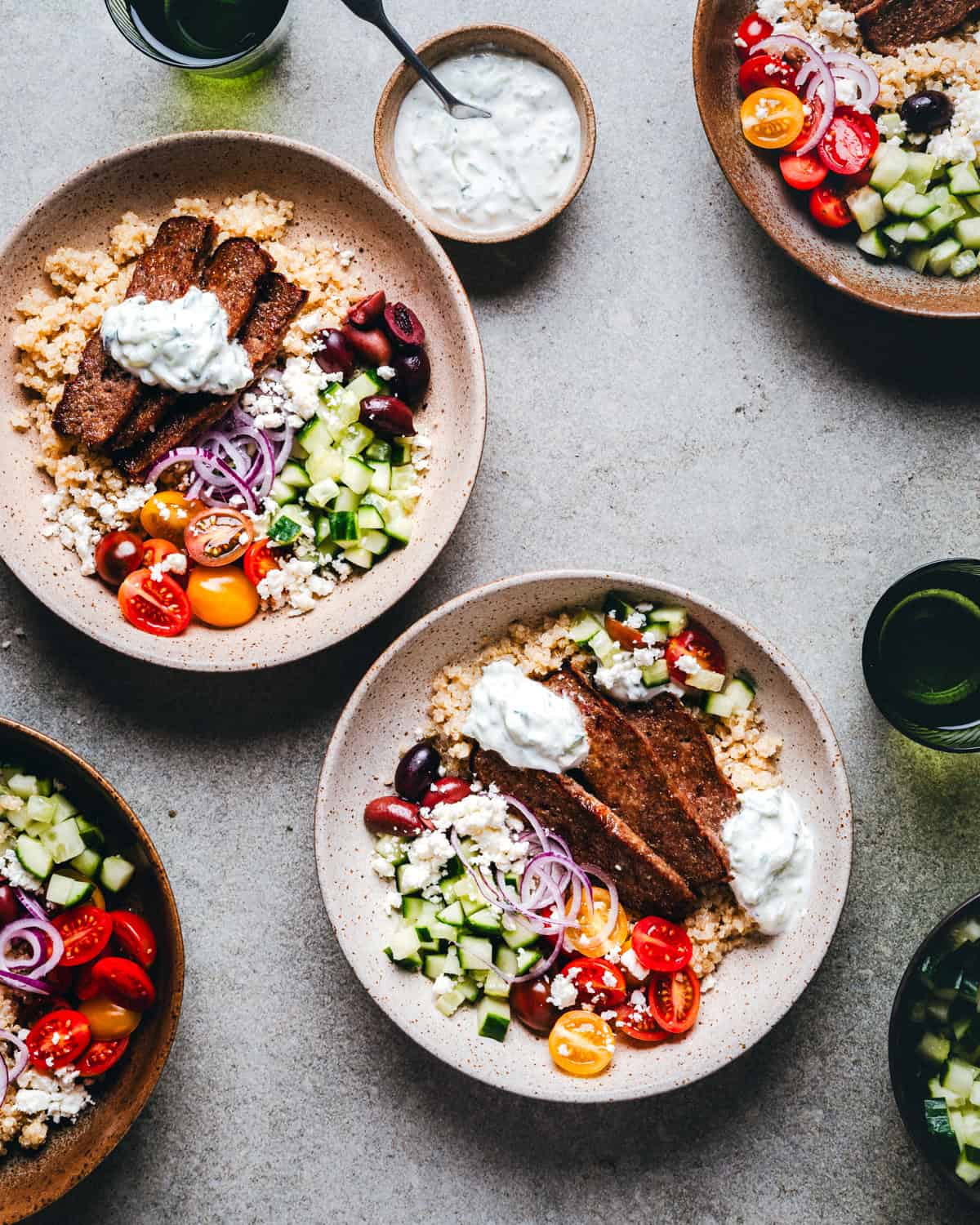 Four gyro bowls with sliced grilled gyro meat, quinoa, cherry tomatoes, cucumber, red onion, olives, and tzatziki sauce. There's a bowl of extra tzatziki and two glasses of water in the background on a gray surface.