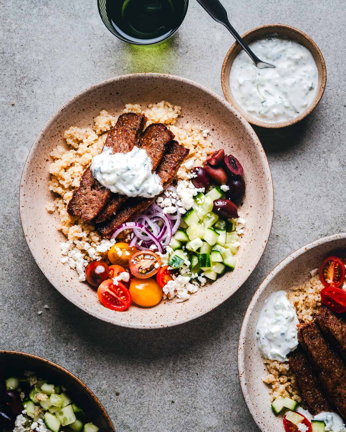 A bowl of couscous topped with grilled gyro meat, cherry tomatoes, cucumber, olives, red onion, and crumbled cheese. A tzatziki sauce is on top. Nearby are a bowl of sauce, a glass of dark liquid, and another similar bowl.