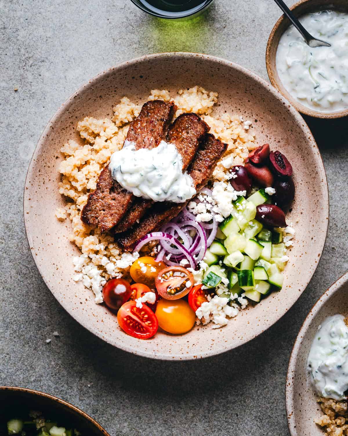 A gyro bowl with couscous, gyro meat slices topped with tzatziki sauce, sliced red onions, cherry tomatoes, cucumber, olives, and crumbled feta cheese on a light-colored surface.