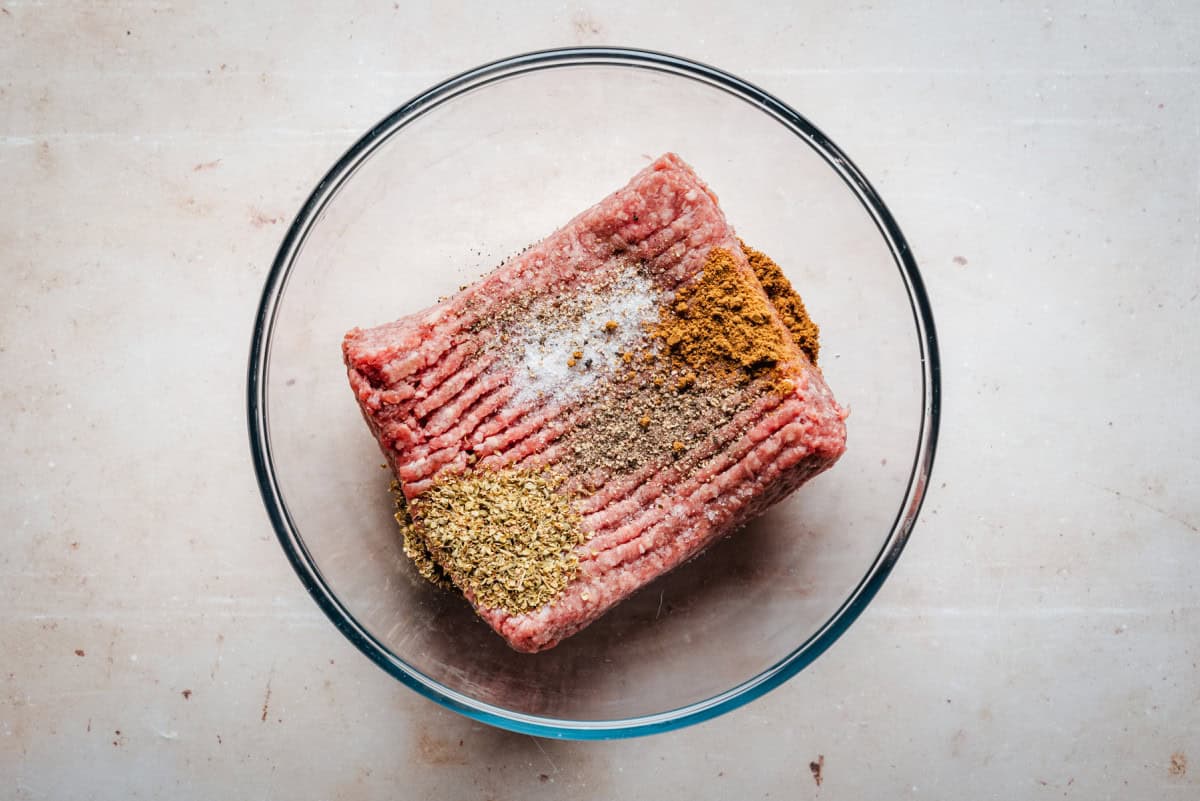 A clear glass bowl containing raw ground lamb sprinkled with spices sits on a beige countertop.
