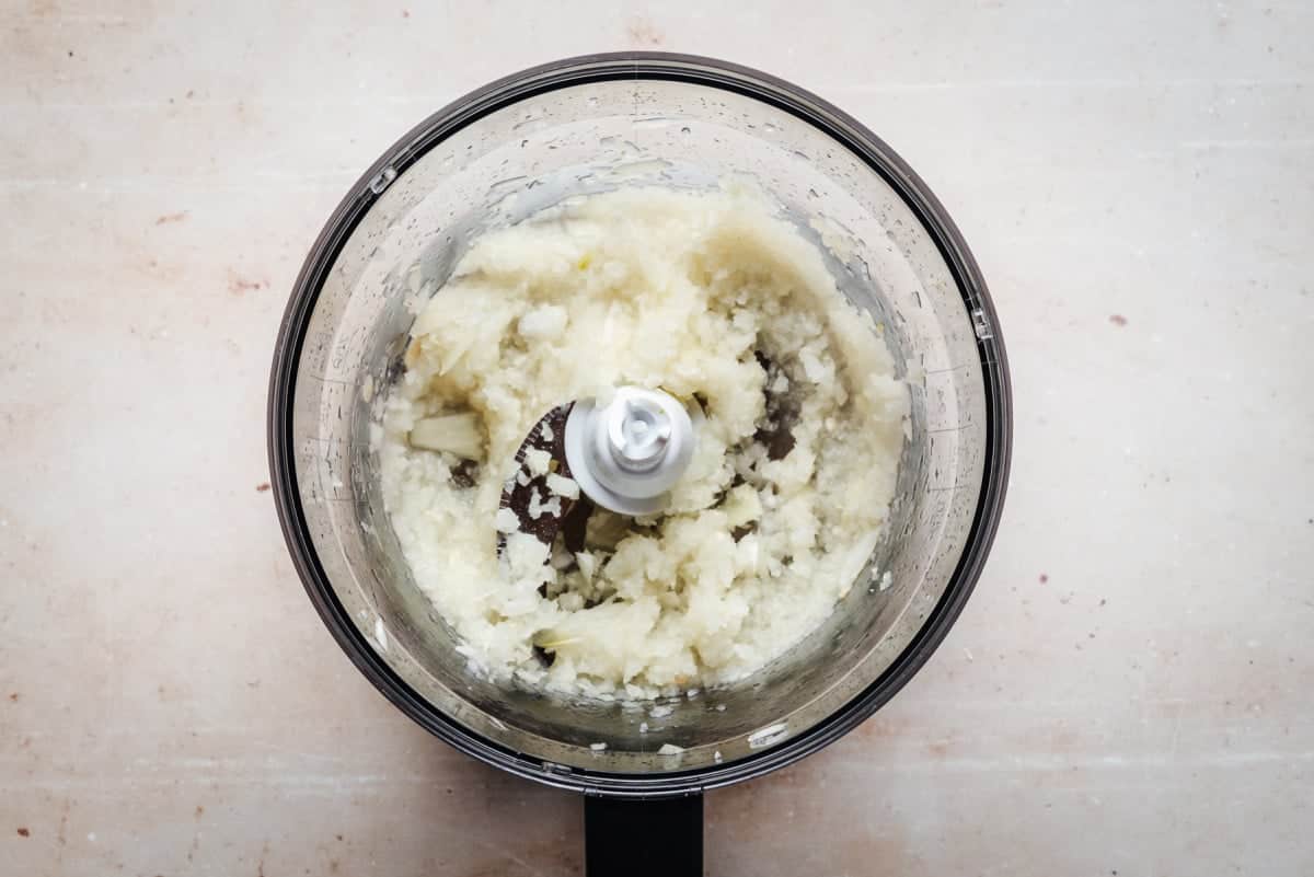 Overhead view of a food processor containing finely chopped or pureed onions, against a light stone background.