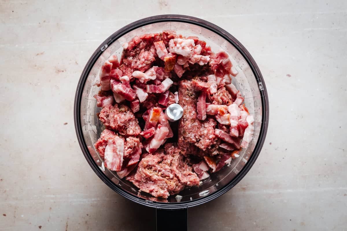 A food processor viewed from above, filled with chunks of raw lamb and bacon. The background is a light-colored countertop.