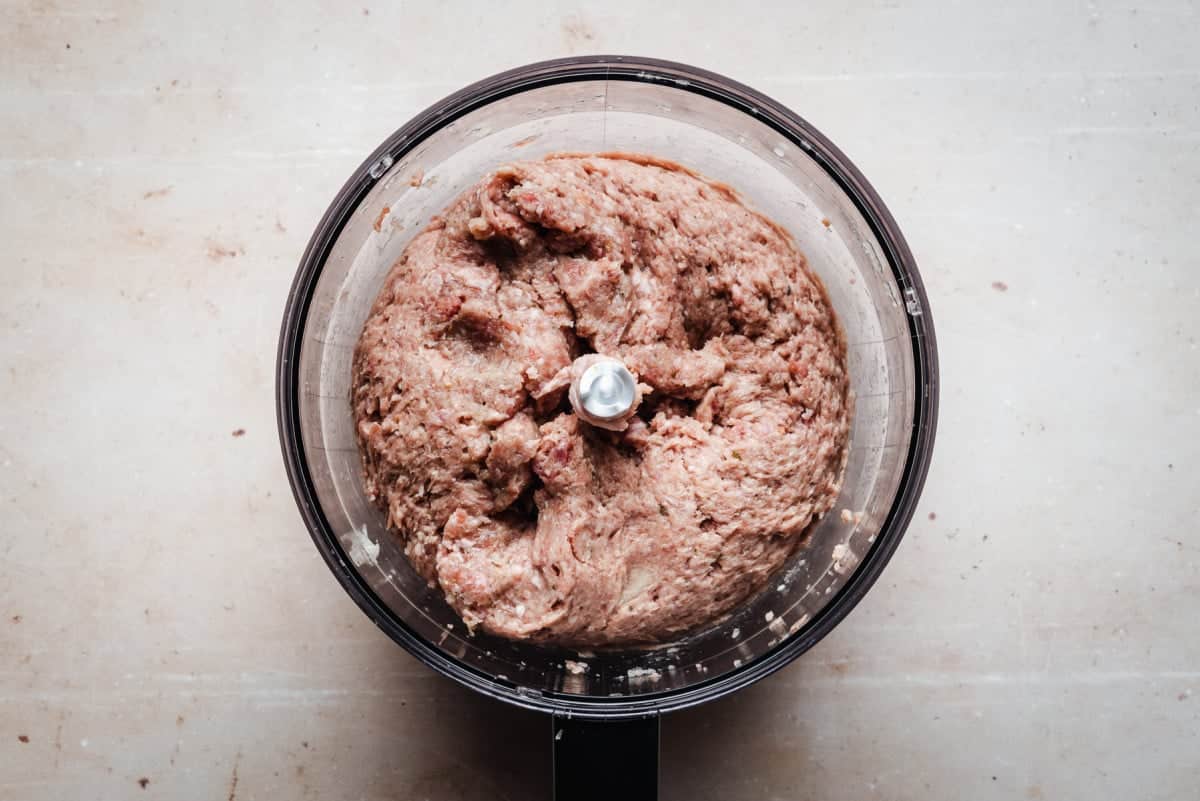 A top-down view of a food processor containing mixed ground meat on a light-colored counter. The meat mixture is pinkish-brown, and the metal blade is visible in the center.