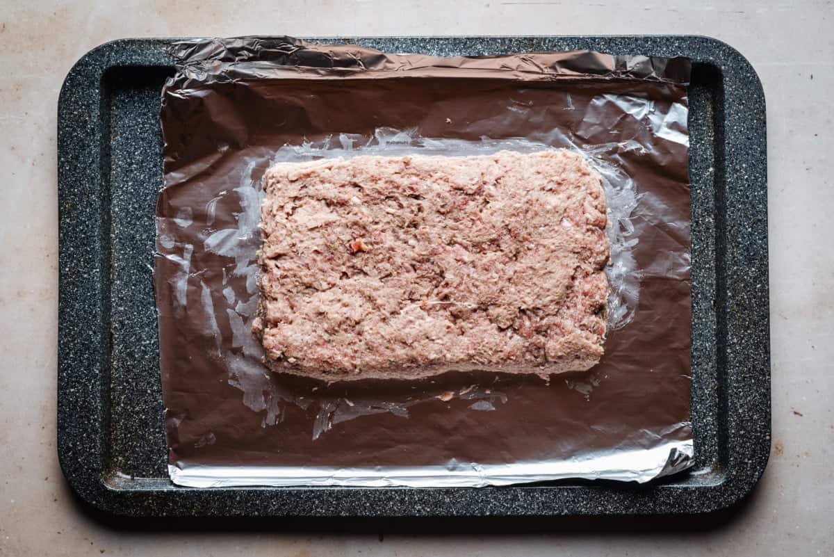 A rectangular raw gyro meat mixture is placed on a foil-lined baking sheet. The baking sheet is dark and textured, contrasting with the shiny foil.