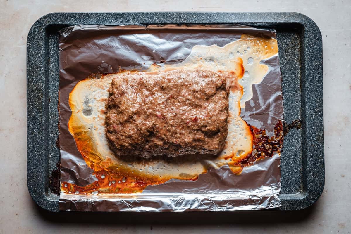 A rectangular loaf of gyro meat sits on aluminum foil atop a dark baking sheet. The meat is surrounded by bubbly juices and lightly browned edges, indicating it has been recently baked.