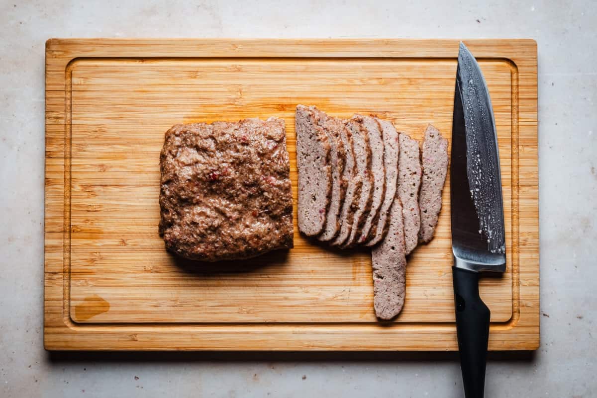 A partially sliced loaf of gyro meat sits on a wooden cutting board. Several slices are neatly arranged to the right. A large kitchen knife with a black handle is placed beside.