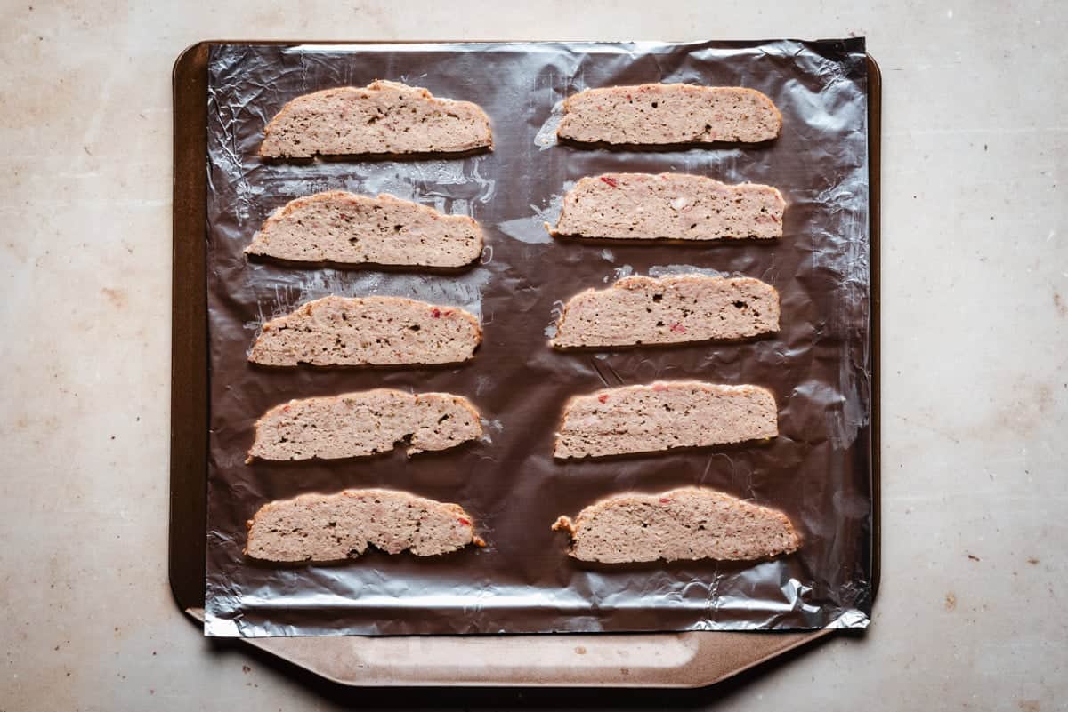 Slices of light brow gyro meat arranged in two columns on a foil-lined baking tray. The surface appears textured with small bits of seasoning. The tray is placed on a light marble countertop.