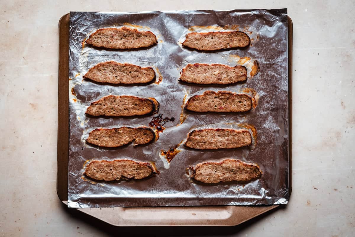 Slices of cooked gyro meat arranged in rows on a foil-lined baking sheet. The slices are evenly spaced, showing a browned outside with visible juices and crumbs on the foil. The background is a neutral countertop.