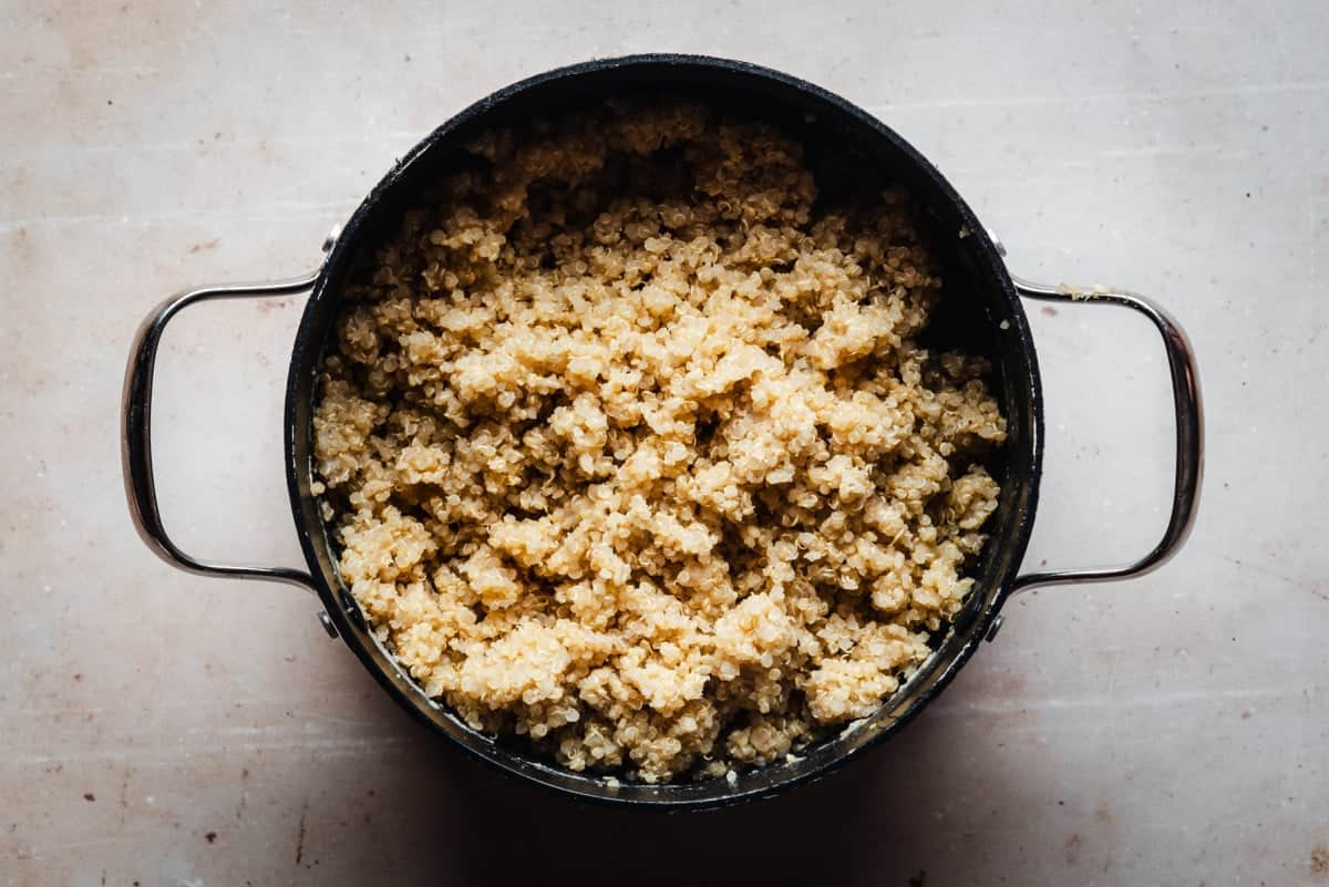 A black pot filled with cooked quinoa, visible from above. The quinoa is light brown and fluffy, with a slightly textured appearance. The pot has metal handles on both sides and rests on a light-colored surface.
