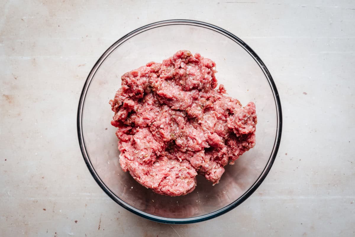 A glass bowl filled with raw ground lamb sits on a light-colored surface.