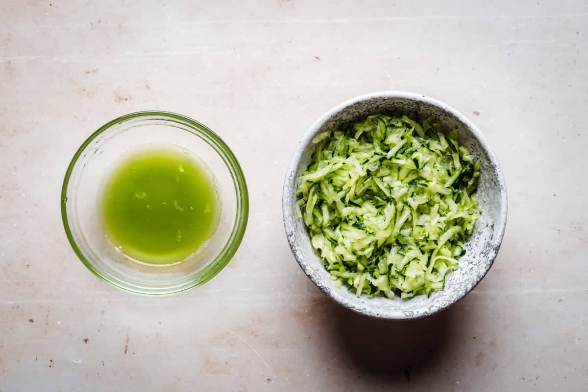 A white bowl filled with shredded cucumber next to a glass bowl containing green cucumber juice. Both are placed on a light-colored surface.