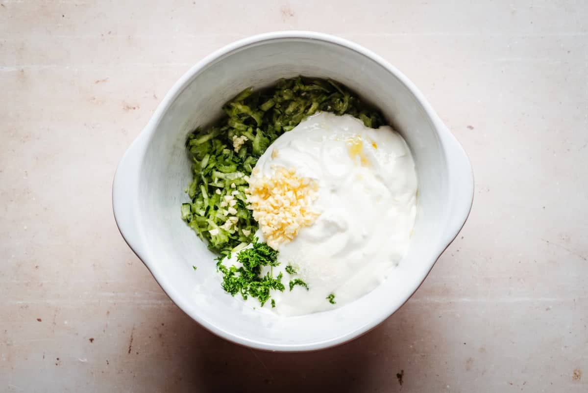 A white bowl contains plain yogurt, grated cucumber and minced dill, ready to be mixed into a sauce. The bowl is set against a light-colored background.