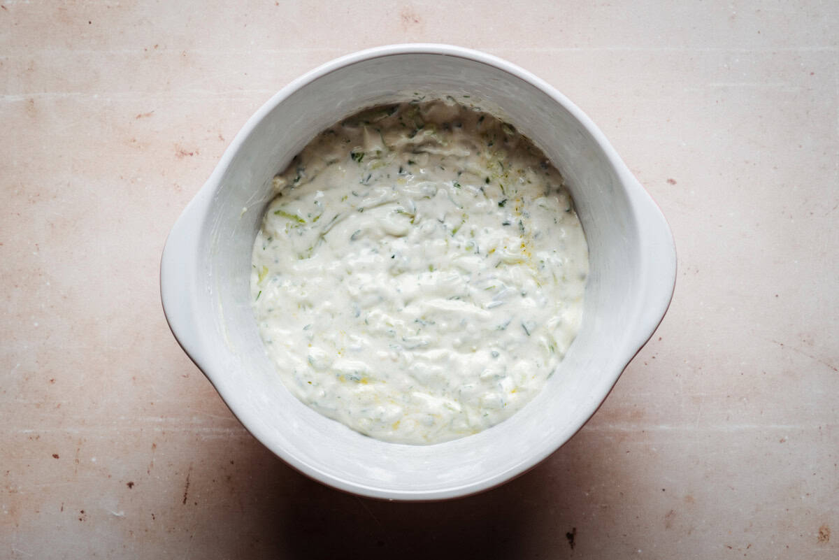 A white bowl filled with creamy tzatziki sauce, featuring visible shreds of cucumber and specks of herbs, sits on a marbled surface. The mixture appears thick and textured.
