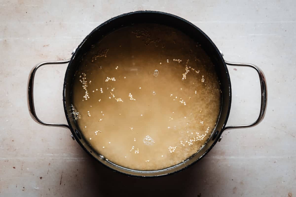 A black pot filled with quinoa grains soaking in water is placed on a light countertop. The water is cloudy, and the grains are visible throughout, creating a textured surface. The pot has two silver handles on each side.