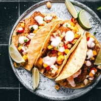 A plate with three fish tacos filled with colorful diced vegetables, grilled fish, and drizzled with white sauce. Lime wedges are placed around the tacos on a textured, dark surface.