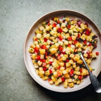 A white bowl filled with colorful diced fruits on a textured surface. The mix includes yellow pineapple, red peppers, and green herbs. A spoon rests on the side of the bowl.