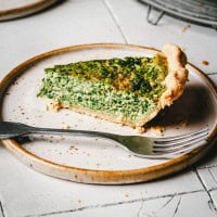 A slice of spinach quiche sits on a ceramic plate, accompanied by a fork. The quiche has a golden-brown crust and a dense, green filling, set on a light-colored, textured surface.