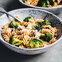 A bowl of fusilli pasta mixed with broccoli florets and ground meat, topped with grated Parmesan cheese. A fork is resting in the bowl. The bowl is on a textured gray surface, and a second similar bowl is partially visible in the background.
