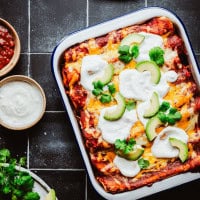 A dish of shredded beef enchiladas topped with melted cheese, avocado slices, and cilantro is placed on a black tiled surface. Beside it, small bowls contain salsa and sour cream, with fresh cilantro and lime wedges on a plate nearby.