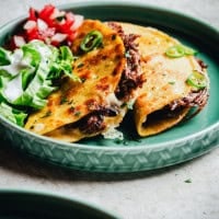 A close-up of two crispy tacos filled with shredded beef and garnished with slices of green chili on a green plate. Next to them are fresh lettuce, sour cream, and chopped tomatoes.
