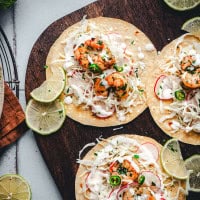 Three spicy shrimp tacos topped with chopped herbs, sliced jalapeños, shredded cabbage, radish slices, and creamy sauce are served on a wooden board. Lime slices are placed around the tacos for garnish.