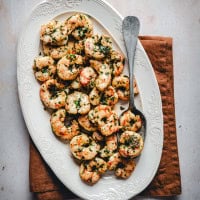 An oval white plate filled with cooked spicy shrimp garnished with chopped herbs and spices, resting on a folded brown napkin. A simple metal serving spoon is placed on the side of the plate. The table surface is off-white.