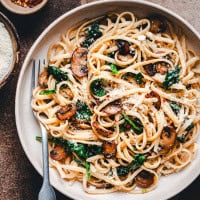 A bowl of Spinach Mushroom Pasta with sautéed mushrooms, spinach, and grated cheese. The dish is garnished with red pepper flakes and is accompanied by a fork. Small bowls with grated cheese and red pepper flakes are on the side.