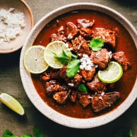 A bowl of Carne Adovada garnished with lime slices, chopped onions, and cilantro. A small dish with more chopped onions and a lime wedge are on the side, all set on a dark green fabric background.