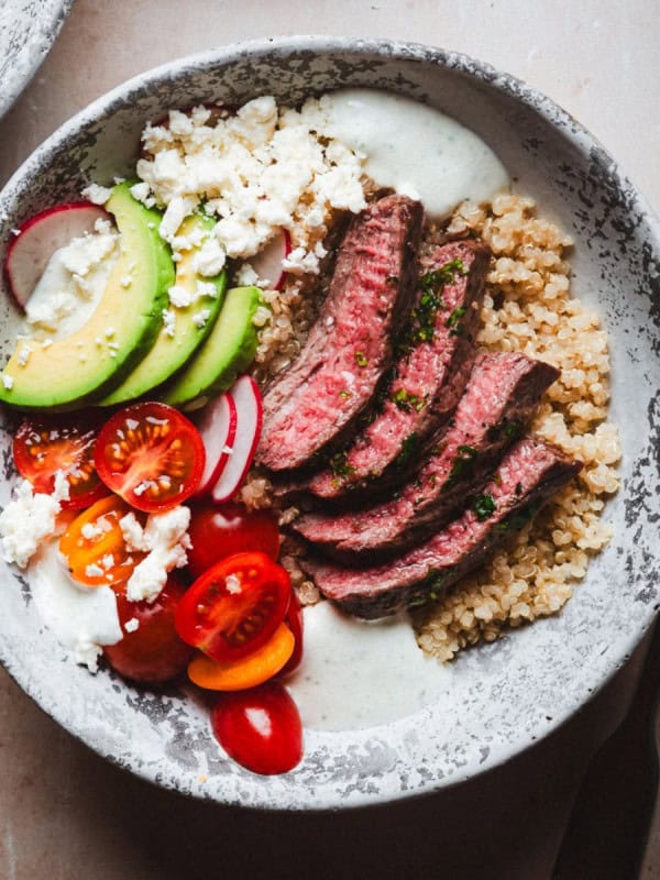 A bowl of quinoa topped with sliced carne asada steak, avocado, cherry tomatoes, radishes, crumbled feta cheese, and a dollop of creamy dressing. A fork lies beside the bowl on a textured surface.