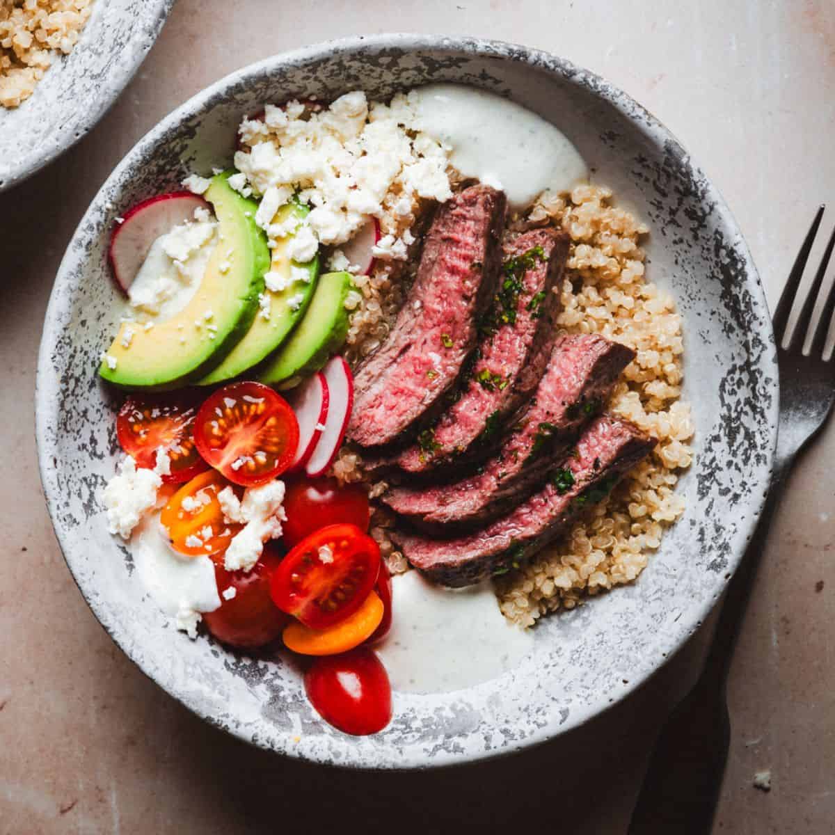 A bowl of quinoa topped with sliced carne asada steak, avocado, cherry tomatoes, radishes, crumbled feta cheese, and a dollop of creamy dressing. A fork lies beside the bowl on a textured surface.