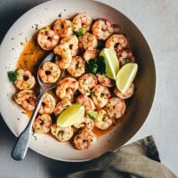 A bowl of cooked cilantro lime shrimp garnished with chopped herbs and lime wedges, with a spoon on the side. The shrimp are arranged in a circular pattern, and the bowl sits on a light gray surface with a dark green napkin nearby.