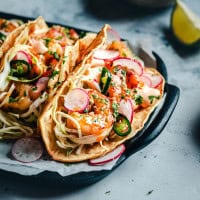 Close-up of three shrimp tacos in a dark dish. The tacos are filled with shrimp, shredded cabbage, sliced radishes, jalapeños, cilantro, and a drizzle of creamy sauce, all on soft tortillas. A lime wedge is in the background.